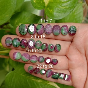 May include: A hand displays a collection of oval and square gemstones in shades of green and red. The gemstones are arranged in rows, with text indicating the quantity of each stone. The background features green foliage.
