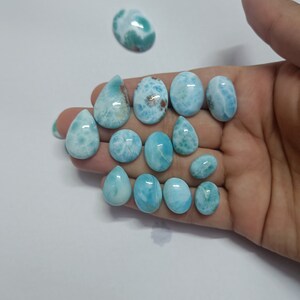 Op de afbeelding: Een verzameling gepolijste larimar edelstenen in ovale en druppelvormige uitvoeringen. De stenen vertonen een reeks lichtblauwe en witte patronen, sommige hebben bruine insluitsels. De edelstenen worden gepresenteerd op een wit oppervlak. Afmetingen in cm.