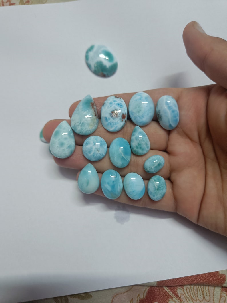 Op de afbeelding: Een verzameling gepolijste larimar edelstenen in ovale en druppelvormige uitvoeringen. De stenen vertonen een reeks lichtblauwe en witte patronen, sommige hebben bruine insluitsels. De edelstenen worden gepresenteerd op een wit oppervlak. Afmetingen in cm.