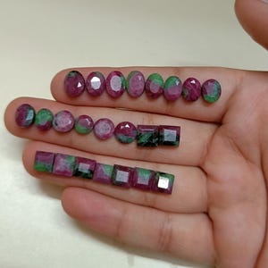 May include: Various ruby gemstones in oval, round, and square shapes. The gemstones display a mix of red and green colors. The gemstones are arranged on a hand, showcasing their different cuts and sizes.
