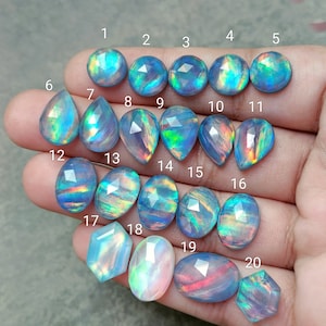 Op de afbeelding: Twintig regenboogkleurige opaal edelstenen in verschillende vormen en maten, genummerd van 1 tot 20. De stenen zijn een mix van blauwe, groene en paarse tinten.