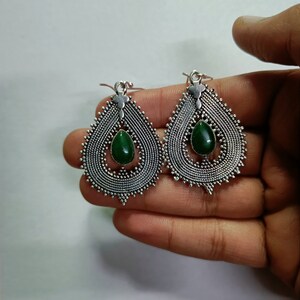 May include: A pair of teardrop-shaped earrings featuring an intricate silver-toned metal filigree frame with a textured, beaded edge. Each earring is centered with a smooth, polished dark green stone in a matching teardrop shape and a hook.