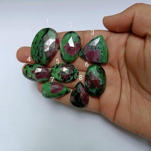 May include: Eight green and pink gemstone cabochons with faceted edges. The stones are arranged in a semi-circle on a hand.