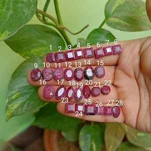 Op de afbeelding: Een verzameling van 28 roze robijn edelstenen in verschillende vormen en maten, waaronder vierkant, ovaal en rond. De edelstenen zijn genummerd van 1 tot en met 28.