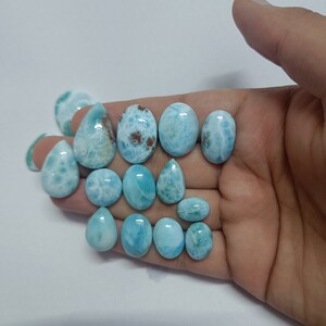 Op de afbeelding: Een verzameling gepolijste larimar edelstenen in diverse vormen, waaronder ovaal en druppelvormig. De stenen vertonen een scala aan blauwtinten met witte en bruine patronen. De edelstenen worden in een hand tegen een witte achtergrond gehouden.