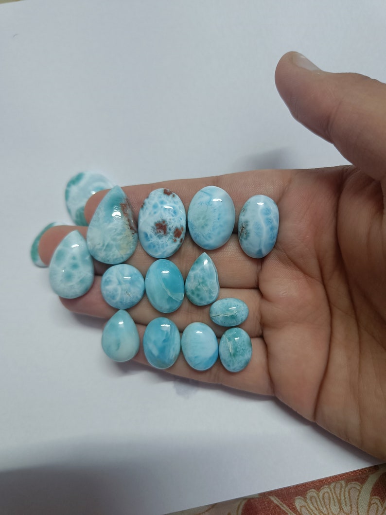 Op de afbeelding: Een verzameling gepolijste larimar edelstenen in diverse vormen, waaronder ovaal en druppelvormig. De stenen vertonen een scala aan blauwtinten met witte en bruine patronen. De edelstenen worden in een hand tegen een witte achtergrond gehouden.