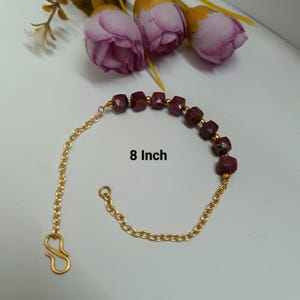 May include: A gold-colored chain bracelet with a series of dark red, square-cut gemstones. The bracelet is 8 inches long and has a clasp closure. The background includes purple flowers.