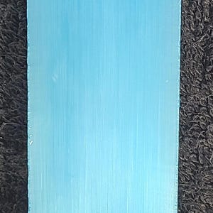 May include: A rectangular, light blue object with a textured surface. The object is set against a dark, textured background. The color is a solid, pale blue.
