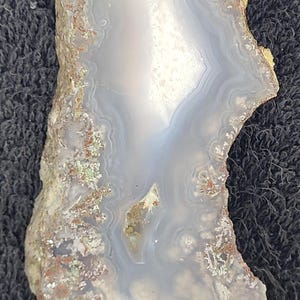 May include: A polished agate slice with a natural, organic shape. The stone features concentric bands of pale blue, white, and translucent areas, with a rough, textured edge. Earthy tones of brown and green are visible.