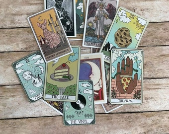 May include: A collection of tarot cards with various illustrations, including a cat, a cake, a pizza, a vinyl record, and a cookie. The cards are laid out on a wooden surface.