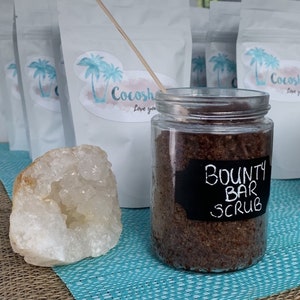May include: A glass jar filled with brown sugar scrub labelled "Bounty Bar Scrub". The jar is sitting on a blue and white patterned surface. There are white bags with a palm tree logo and the text "Cocoshe Love you" in the background.