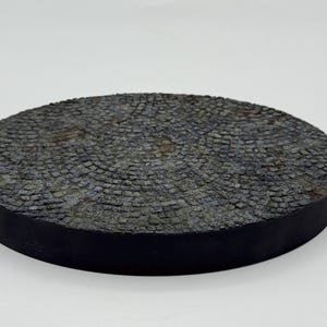 May include: A round, textured, dark gray decorative base with a black edge. The surface has a cobblestone pattern, creating a rustic appearance. The base is elevated and appears to be made of a solid material.