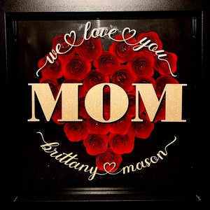 May include: A square black shadow box frame containing a heart-shaped arrangement of red paper roses. The gold glitter text reads "we love you MOM" above the heart and "brittany" and "mason" separated by a small heart icon below the heart.