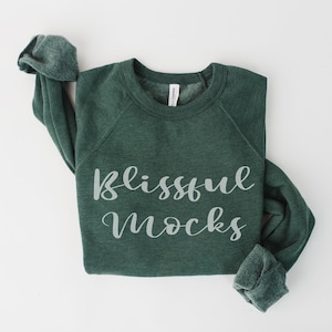 以下が含まれることがあります： 筆記体フォントで「Blissful Mocks」と書かれた、濃い緑色のスウェットシャツ。長袖は折り返されています。スウェットシャツは柔らかい素材でできており、白い背景に表示されています。