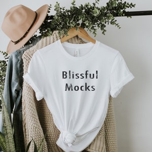 May include: White t-shirt with the text "Blissful Mocks" printed on the front. The shirt is hanging on a wooden hanger and is styled with a brown hat and a beige cardigan.
