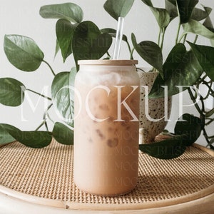May include: A glass of iced coffee with a straw in a clear glass jar on a woven wicker table with a potted plant in the background.