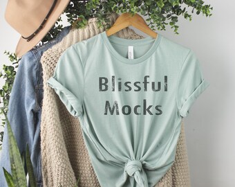 May include: A light green t-shirt with the text "Blissful Mocks" printed on the front. The shirt is tied at the waist and is hanging on a wooden hanger.