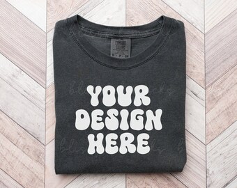 May include: A black t-shirt with the text "YOUR DESIGN HERE" in white, retro-style lettering. The shirt is folded and laid flat on a wooden surface.