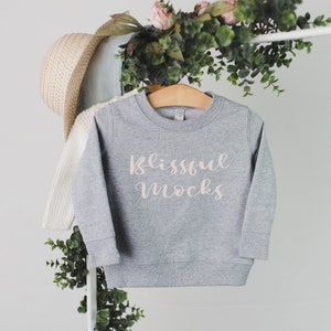 May include: A gray sweatshirt with the text "Blissful Mocks" in a cursive font. The sweatshirt is hanging on a wooden hanger. A straw hat and a white sweater are visible in the background. Greenery and pink flowers are also present.