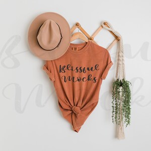 May include: A rust-colored t-shirt with the text "Blissful Mocks" printed on the front. The t-shirt is hanging on a wooden rack with a straw hat and a macrame plant hanger.