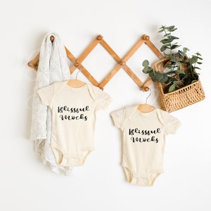 May include: Two cream-colored baby onesies hanging on a wooden rack. Both onesies have the text "Blissful mocks" printed in black.