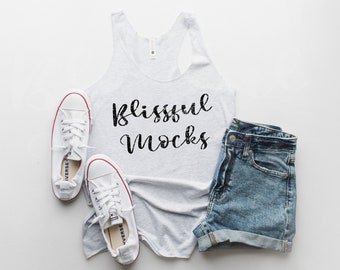 May include: A white tank top with the text "blissful mocks" in black lettering. The tank top is paired with denim shorts and white trainers.