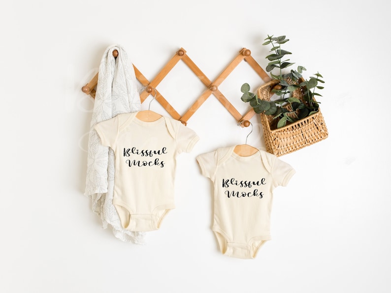 May include: Two cream-colored baby onesies hanging on a wooden rack. Both onesies have the text "Blissful mocks" printed in black.