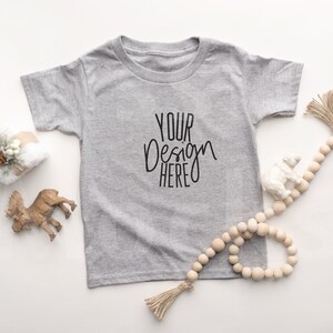 May include: A grey short-sleeved t-shirt with the text "YOUR DESIGN HERE" printed on the front. The t-shirt is lying flat on a white surface with wooden beads, a small wooden reindeer, and a wrapped gift.