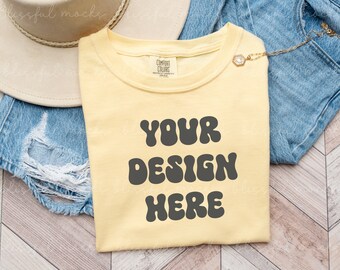 May include: A yellow t-shirt with the text "YOUR DESIGN HERE" in a retro font. The shirt is laying on a wooden surface with a pair of blue jeans and a straw hat.