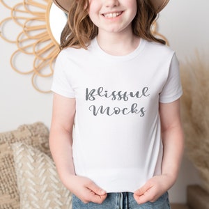May include: A white t-shirt with short sleeves featuring the text "Blissful Mocks" in gray script font. The shirt is being worn by a person with a brown hat and long brown hair.