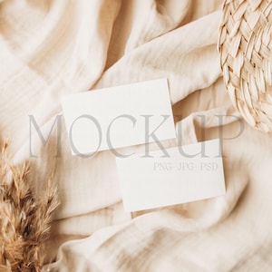 May include: Two blank white business cards on a beige fabric background with a woven straw hat and a dried pampas grass plume.
