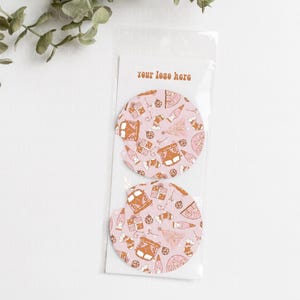 May include: Two pink round coasters with a brown and white pattern of a vintage van, Christmas trees, and other holiday designs. The text "your logo here" is printed on a white card behind the coasters.