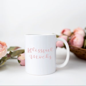 K&ouml;nnte beinhalten: Eine wei&szlig;e Keramiktasse mit einer rosa Aufschrift, die "Blissful Mocks" lautet. Die Tasse steht auf einer wei&szlig;en Oberfl&auml;che mit rosa Blumen im Hintergrund.