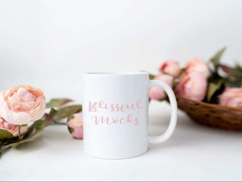 K&ouml;nnte beinhalten: Eine wei&szlig;e Keramiktasse mit einer rosa Aufschrift, die "Blissful Mocks" lautet. Die Tasse steht auf einer wei&szlig;en Oberfl&auml;che mit rosa Blumen im Hintergrund.