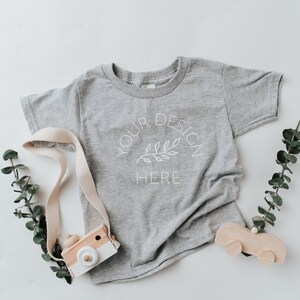 Puede incluir: Una camiseta gris con un dise&ntilde;o blanco que dice "Your Design Here" con una peque&ntilde;a rama y hojas. La camiseta est&aacute; puesta plana sobre una superficie blanca con una c&aacute;mara de juguete de madera y un coche de juguete de madera.