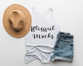 May include: A white tank top with the text "Blissful Mocks" printed on the front. The tank top is lying flat on a white surface with a straw hat and denim shorts beside it.