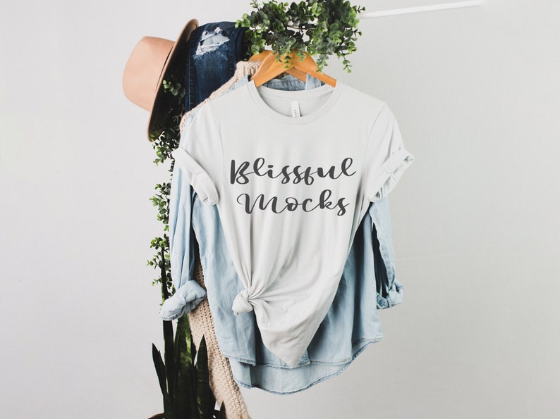 May include: A light gray t-shirt with the text "Blissful Mocks" printed on the front. The shirt is hanging on a wooden hanger and is styled with a denim shirt and a brown hat.