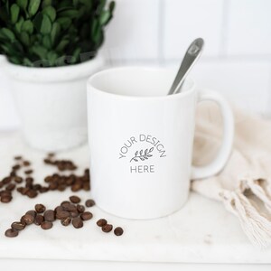 Puede incluir: Taza de cer&aacute;mica blanca con un dise&ntilde;o negro que dice "YOUR DESIGN HERE" con un peque&ntilde;o gr&aacute;fico de rama. La taza est&aacute; sobre una superficie de m&aacute;rmol blanco con granos de caf&eacute; esparcidos a su alrededor.