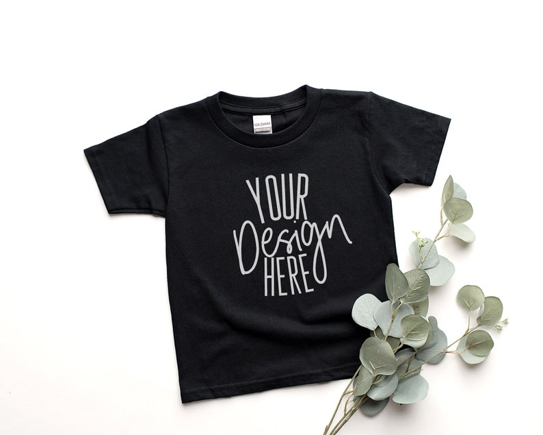 May include: Black t-shirt with the text "YOUR Design HERE" in white script. The short-sleeved shirt is displayed on a white surface, with a sprig of green and grey leaves in the lower right corner.
