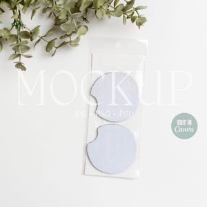 May include: Two white blank round car coasters in a clear plastic package. The package has the text "MOCKUP" and "JPG PNG PSD" printed on it. The text "EDIT IN Canva" is printed on a light blue circle.