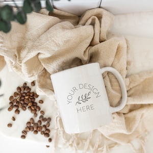 Könnte beinhalten: Eine weiße Keramiktasse mit einem schwarzen Design, auf dem "YOUR DESIGN HERE" steht und ein kleiner Zweig mit Blättern. Die Tasse steht auf einer weißen Oberfläche mit einem beigefarbenen Stoff und verstreuten Kaffeebohnen.