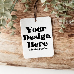 May include: A white rectangular tag with the text "Your Design Here" in a bold, black font, and "Blissful Mocks" below. The tag hangs from a black string, set against a wooden surface with green foliage.