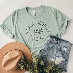 May include: A light green t-shirt with a simple design featuring a small branch with leaves and the text "YOUR DESIGN HERE". The t-shirt is laid flat on a white surface with a brown hat, blue denim shorts, and green foliage.