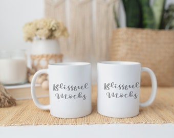 Puede incluir: Dos tazas de cerámica blanca con texto negro que dice "Blissful Mugs". Las tazas están sobre un mantel tejido marrón.