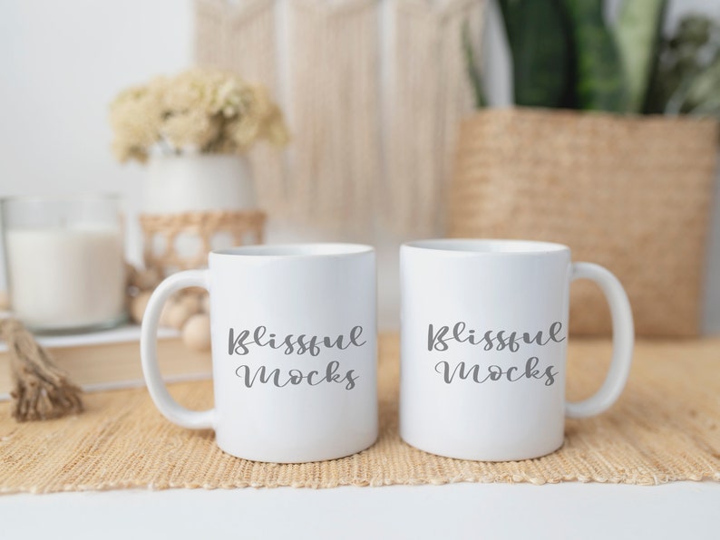 May include: Two white ceramic mugs with black text that reads "Blissful Mugs". The mugs are sitting on a woven brown placemat.