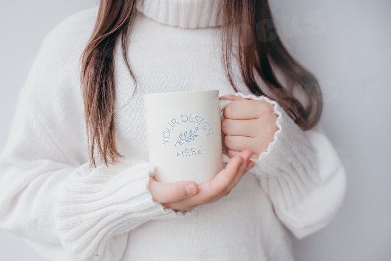 May include: A white ceramic mug with the text "YOUR DESIGN HERE" and a small blue leaf design. The mug is being held by a person wearing a white sweater.