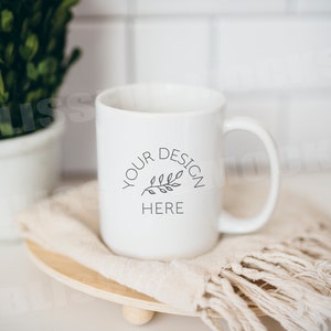 Könnte beinhalten: Weiße Keramiktasse mit einem schwarzen Design, auf dem "YOUR DESIGN HERE" steht und ein kleiner Zweig mit Blättern. Die Tasse steht auf einem weißen und braunen, fransigen Tuch.