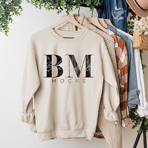 以下が含まれることがあります： 前面に「BM」と「MOCKS」と書かれた黒いグラフィックが施されたベージュのトレーナー。トレーナーは木製ハンガーにかけられています。