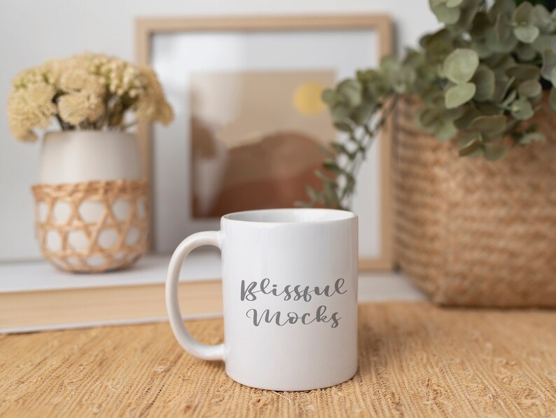 May include: White ceramic mug with a black inscription that reads "Blissful Mocks". The mug is sitting on a woven brown mat.