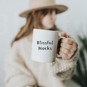Könnte beinhalten: Eine weiße Keramiktasse mit dem Text "Blissful Mocks" in schwarzen Buchstaben. Die Tasse wird von einer Person gehalten, die einen beigen Pullover und einen braunen Hut trägt.